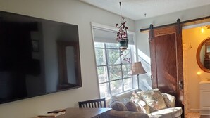 Een woonkamer met een houten eettafel, twee stoelen, een bank en een aan de muur bevestigde televisie.