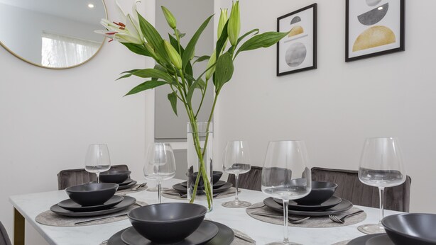Une table à manger dressée avec des bols, des assiettes et des verres à vin noirs, un vase contenant des feuilles vertes et un miroir accroché au mur.