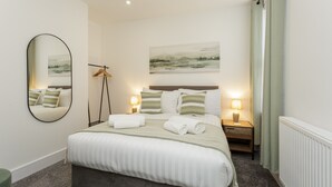 Une chambre à coucher bien rangée, avec un grand lit, deux tables de chevet avec des lampes, un miroir et un tableau accroché au mur.