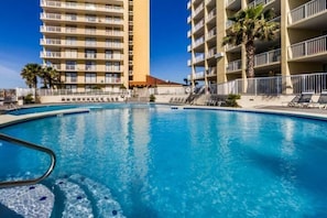 Una piscina con agua cristalina azul, rodeada por una cerca blanca y adyacente a un edificio de varios pisos con balcones.