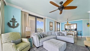 Ein Wohnzimmer mit Ecksofa, einem gemusterten Sessel, einem Deckenventilator und einem großen Fenster mit Vorhängen.