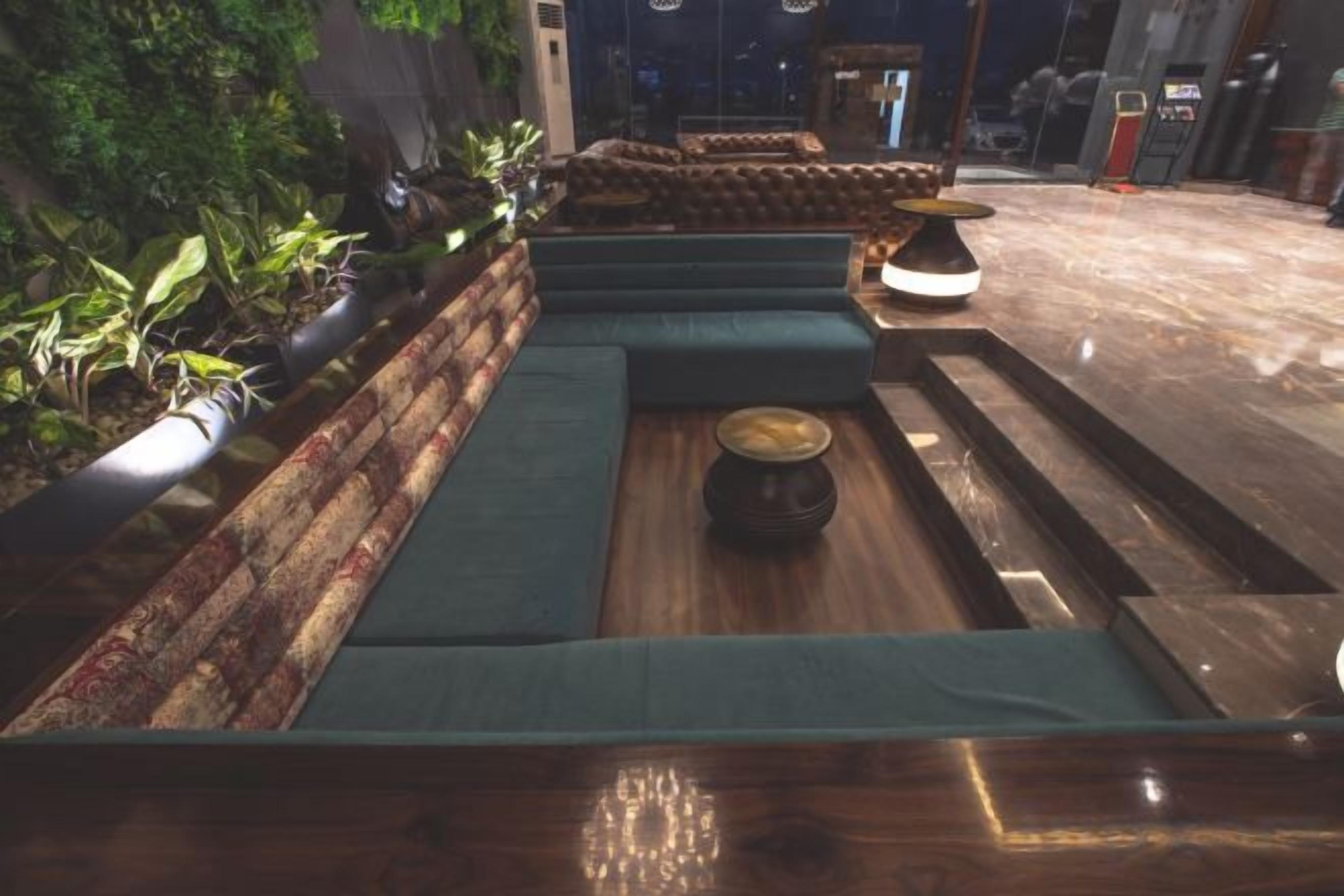 Una zona de estar con bancos acolchados y una mesa central, rodeada de plantas.