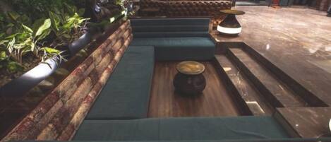 Una zona de estar con bancos acolchados y una mesa central, rodeada de plantas.
