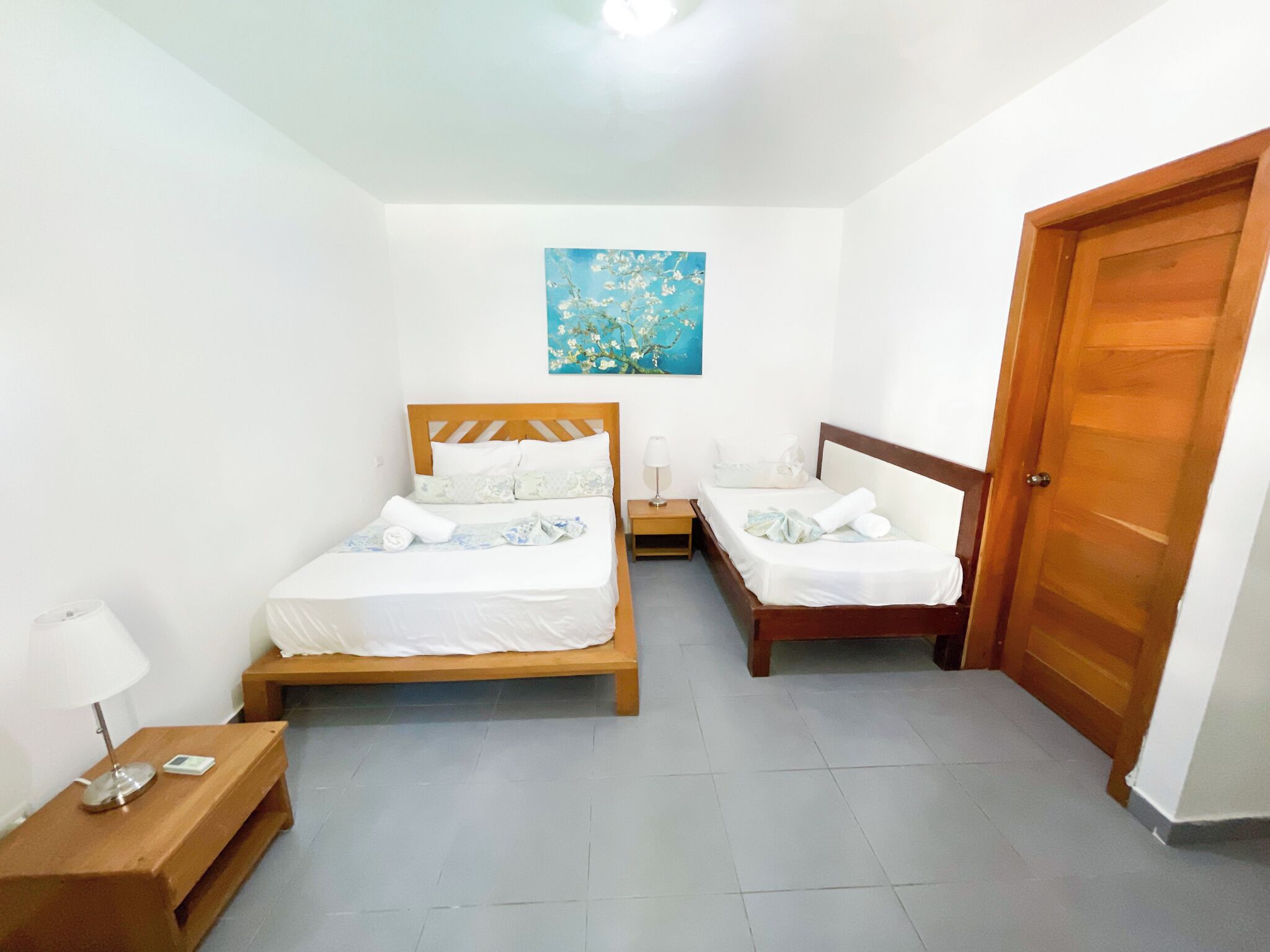 Una habitación pequeña y limpia con dos camas de madera, una mesita de noche con lámpara, un banco de madera y una puerta.