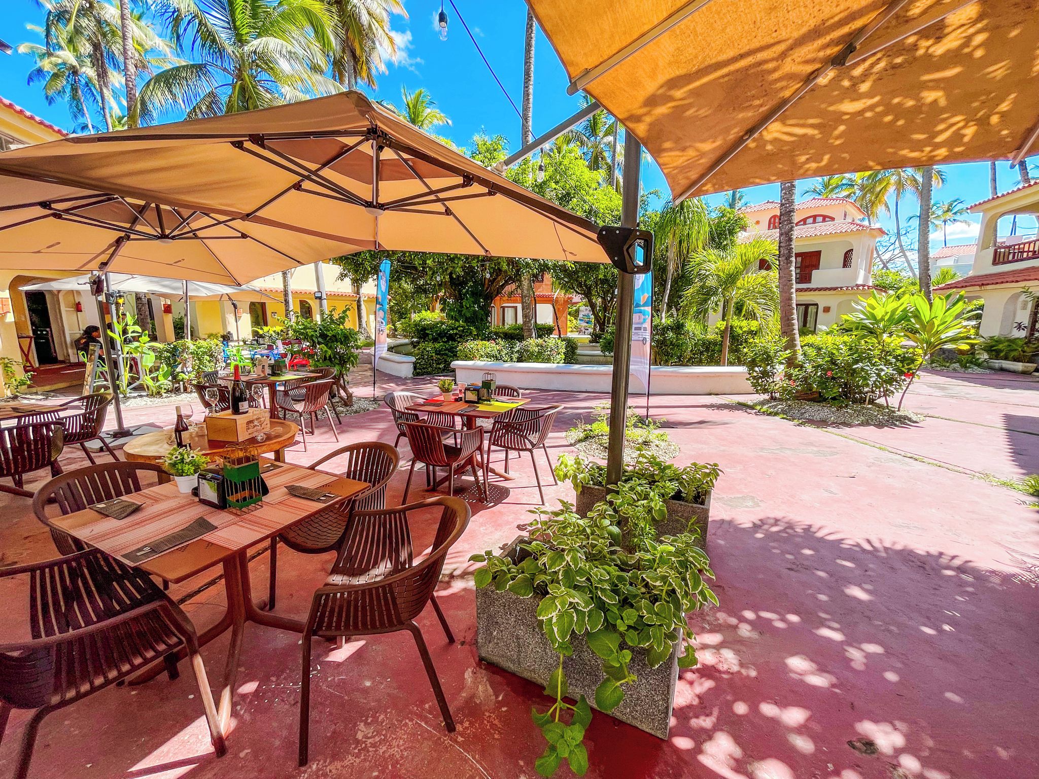 An outdoor café with tables and chairs under canopies, surrounded by greenery and buildings.