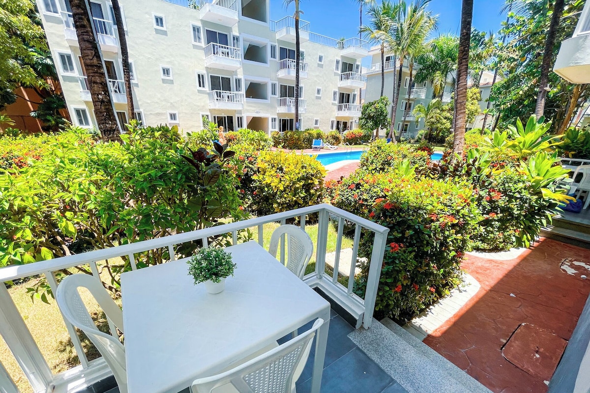 A balcony with a white table and chairs, surrounded by lush greenery and a pool in the background.