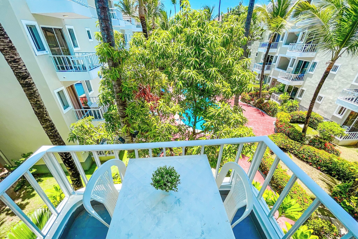 A balcony with a table and chairs, surrounded by lush greenery and a pool.