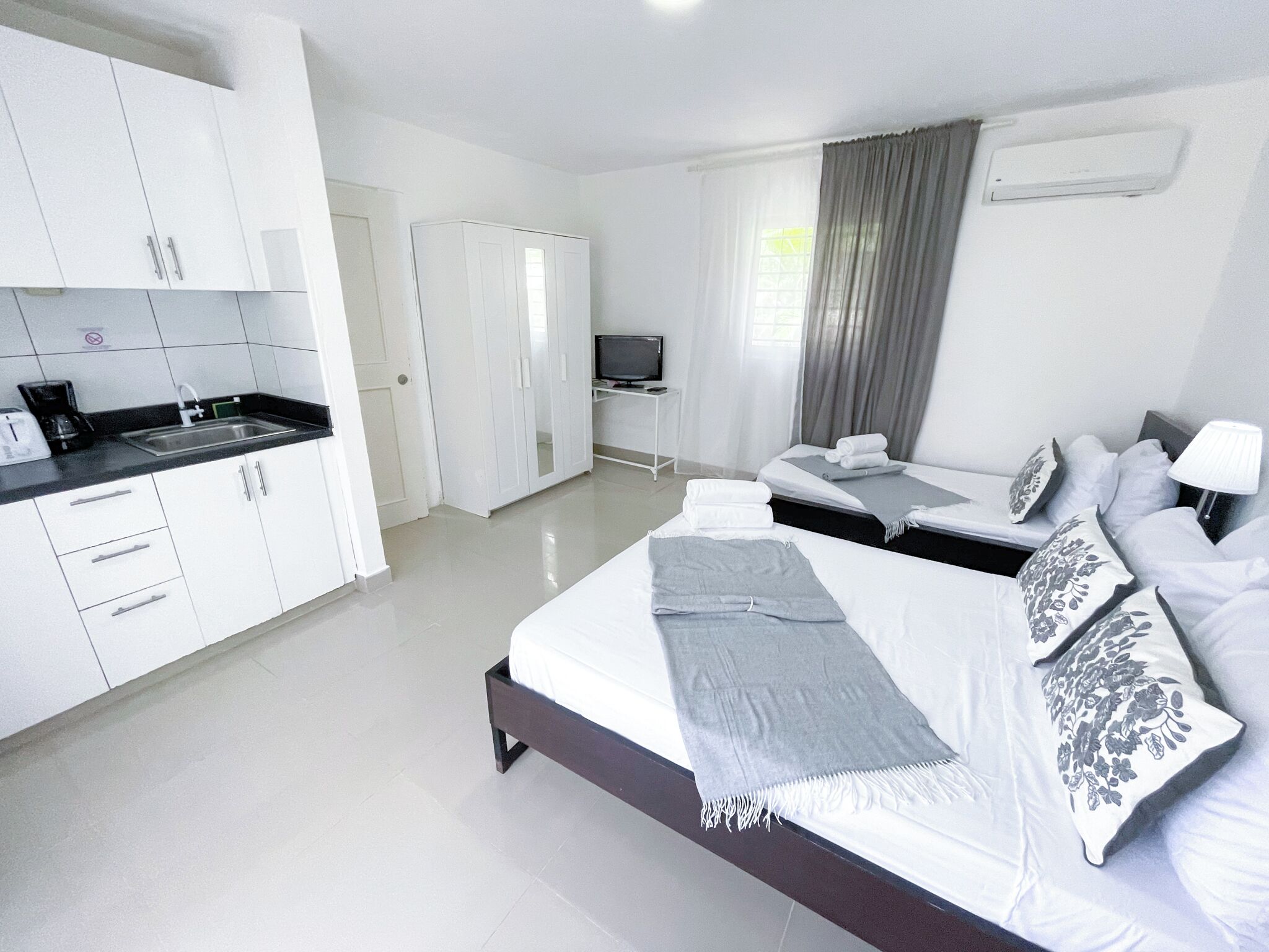 Een moderne hotelkamer met een keukenhoek, een bed met veel kussens, een televisie en een raam met gordijnen.