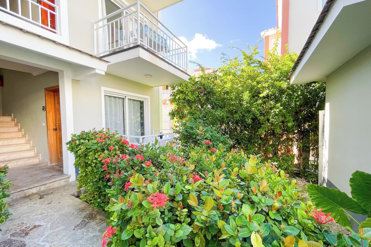 A two-story building with a balcony, a staircase leading to the entrance, and a garden with flowering plants.
