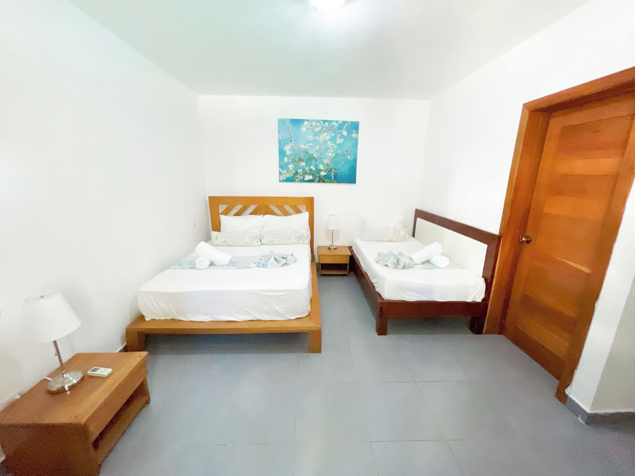 A bedroom with two beds, a wooden door, a nightstand, and a painting on the wall.