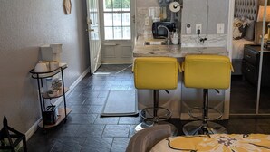 Eine kleine Küche mit Fenster, Spüle und Arbeitsplatte. Ein gelber Stuhl und ein Bett mit Blumenmuster sind ebenfalls vorhanden.