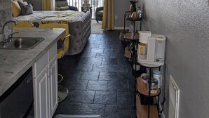 Ein kleines Hotelzimmer mit Küchenzeile, einem Bett mit gemustertem Überwurf, einem Deckenventilator und Blick durch ein großes Fenster nach draußen.