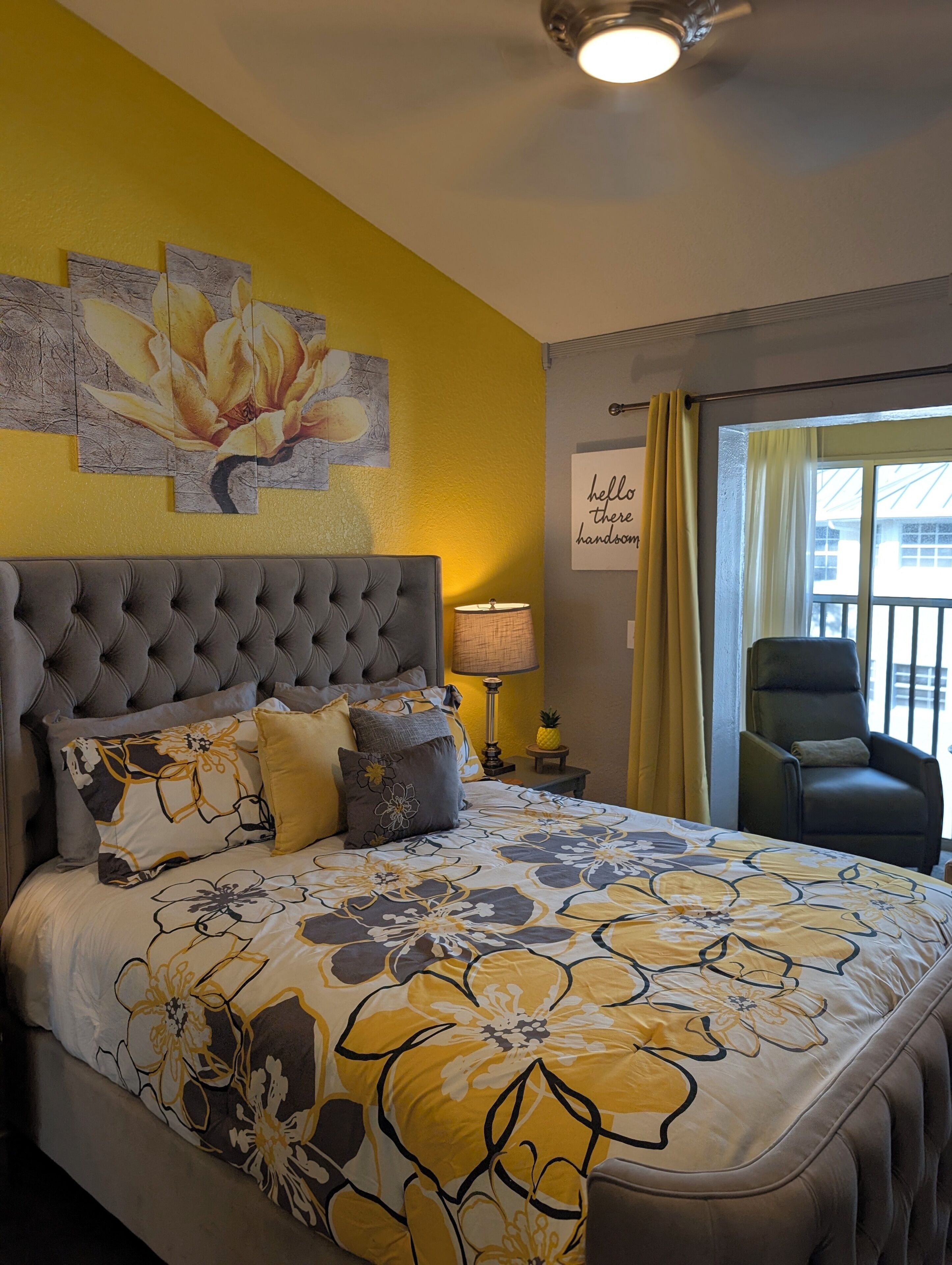 A bedroom with a tufted headboard, floral bedding, a nightstand with a lamp, a chair, and a window with curtains.