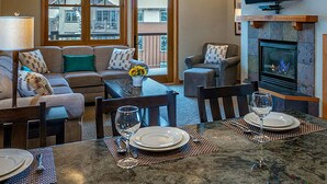 Un salon comprenant un canapé, des fauteuils, une table à manger dressée pour deux personnes, une cheminée et un téléviseur fixé au mur.