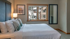 Un lit bien fait, avec du linge de lit blanc et plusieurs oreillers, une lampe de chevet, un tableau encadré au mur, une fenêtre donnant sur des bâtiments et des arbres, et un miroir au mur.