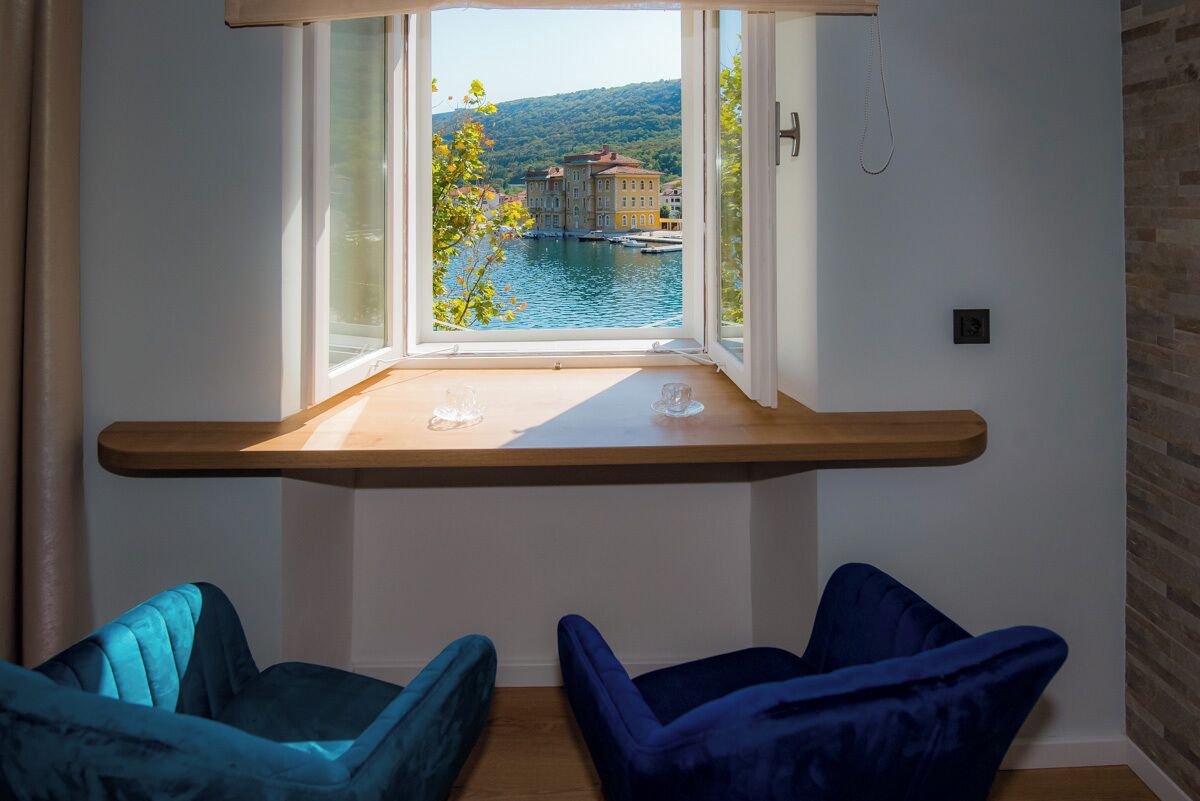A room with a window overlooking a body of water and buildings, a wooden shelf, and two blue chairs.