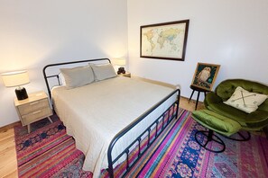 Une chambre à coucher bien rangée, avec un lit, un fauteuil vert, une petite table avec une lampe, une carte du monde encadrée et un tableau représentant une chouette, également encadré.