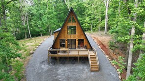 Une cabane en bois avec un toit triangulaire, une terrasse dotée de meubles d’extérieur et une allée de gravier bordée d’arbres.