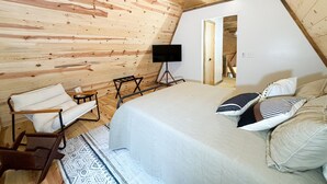 Una camera da letto con soffitto spiovente, un letto con molti cuscini, una sedia, un tavolino e una TV su un mobile.