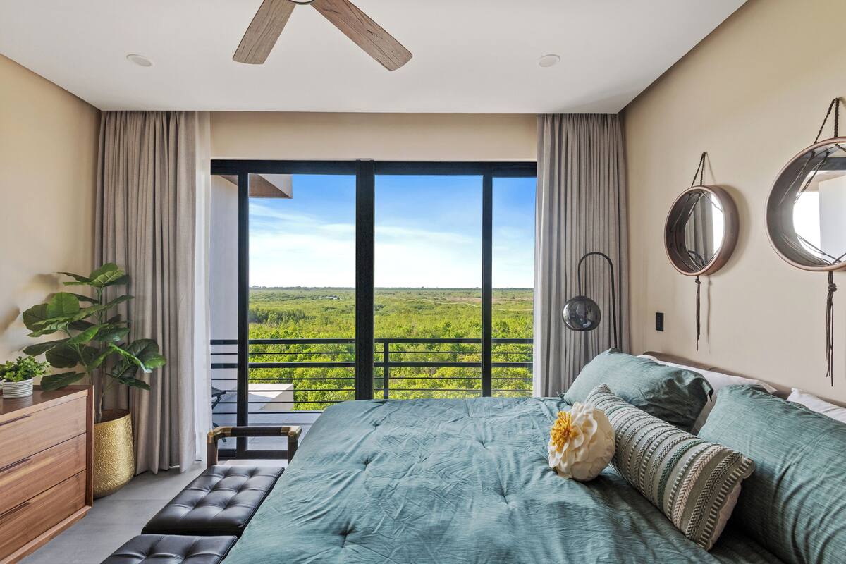 A bedroom with a large bed, a ceiling fan, a view of greenery through a balcony, and a potted plant.