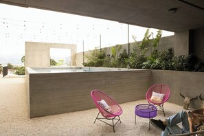 Un área moderna de piscina al aire libre con paredes de concreto, dos sillas de mimbre rosa y una morada, un adorno con forma de cráneo de toro y vistas a la ciudad al fondo.