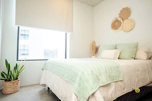 A bedroom with a large window, a bed with a green blanket, a woven plant pot, and decorative wall art. - Catorce Urbano (San José)