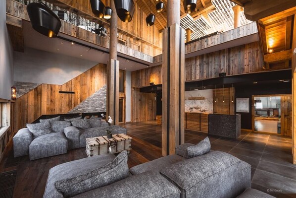 Ein geräumiges Foyer mit hoher Decke, Holzvertäfelung und einer Treppe zu einer oberen Ebene. Es gibt Sitzbereiche mit grau gepolsterten Sofas und einem kleinen Tisch mit einer Topfpflanze.