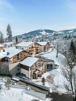 Ein schneebedecktes Chalet mit Holzhütten, einem Steinhaus und einer Skipiste im Hintergrund.