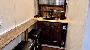 Een kleine keuken met een houten spatwand, een spoelbak en een koffieapparaat. Er zijn twee krukken en een aanrecht.