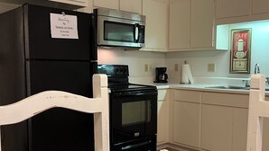 Een keuken met een zwarte koelkast, een magnetron, een fornuis en een koffiezetapparaat. Er staat een witte stoel op de voorgrond en aan de muur hangt een lijst met een afbeelding.