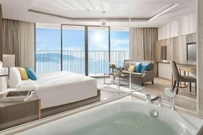 A modern hotel room with a large bed, a sitting area with a coffee table, a desk with a chair, a large window with a view of the ocean, and a bathtub. - Panorama Nha Trang SanVilla (Nha Trang)