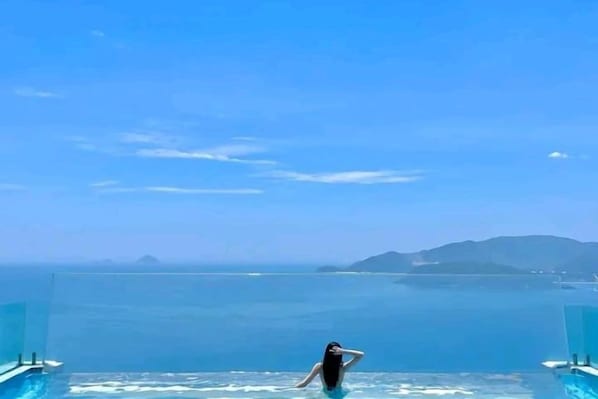 Une personne se tient au bord d’une piscine, d’où l’on aperçoit l’océan et la côte au loin.