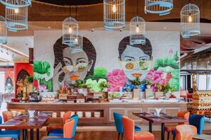 Een restaurant met een muurschildering, hangende lampen en kleurrijke zitplaatsen.