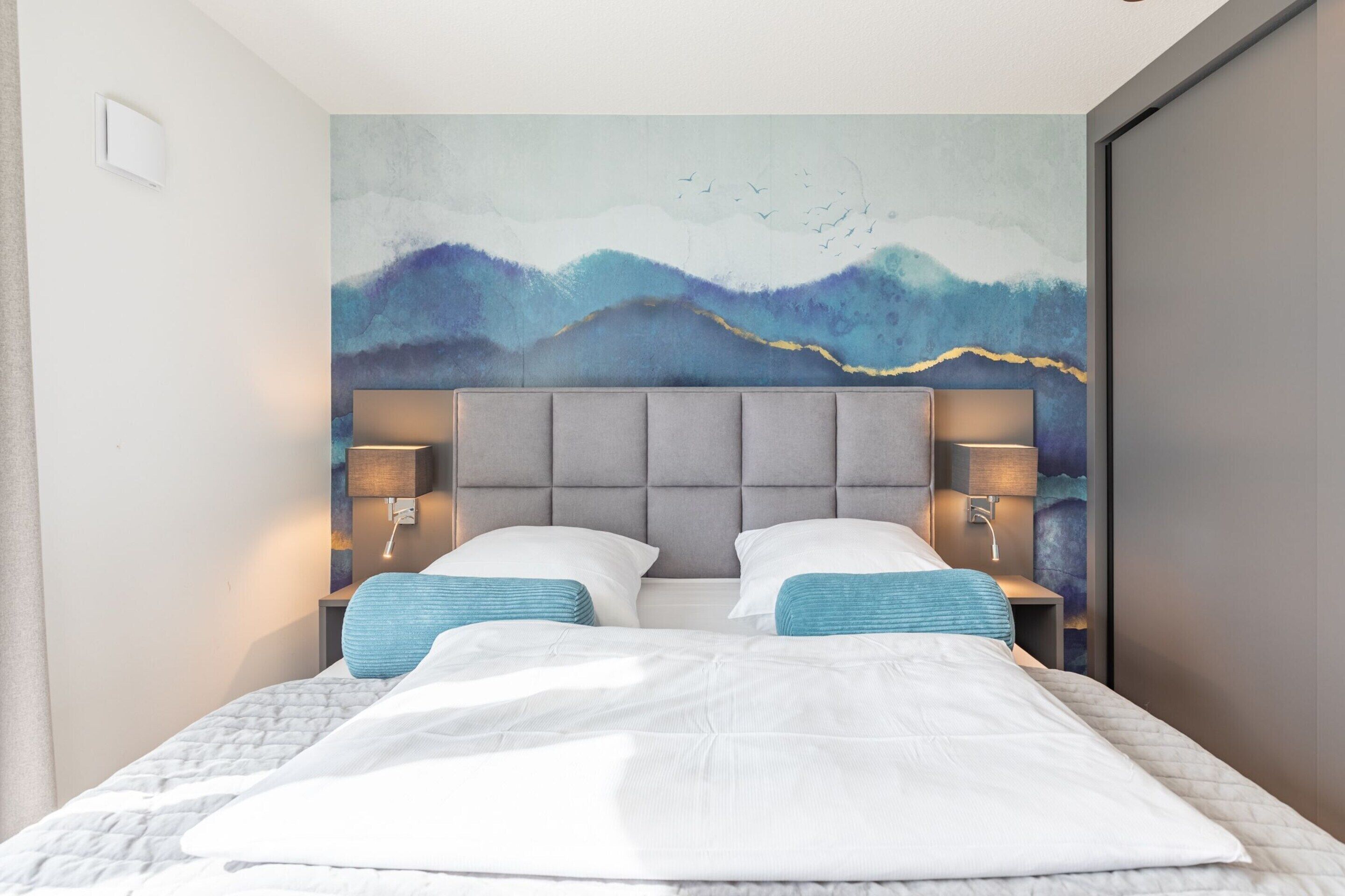 A modern hotel room with a large bed, two bedside tables with lamps, and a wall mural.
