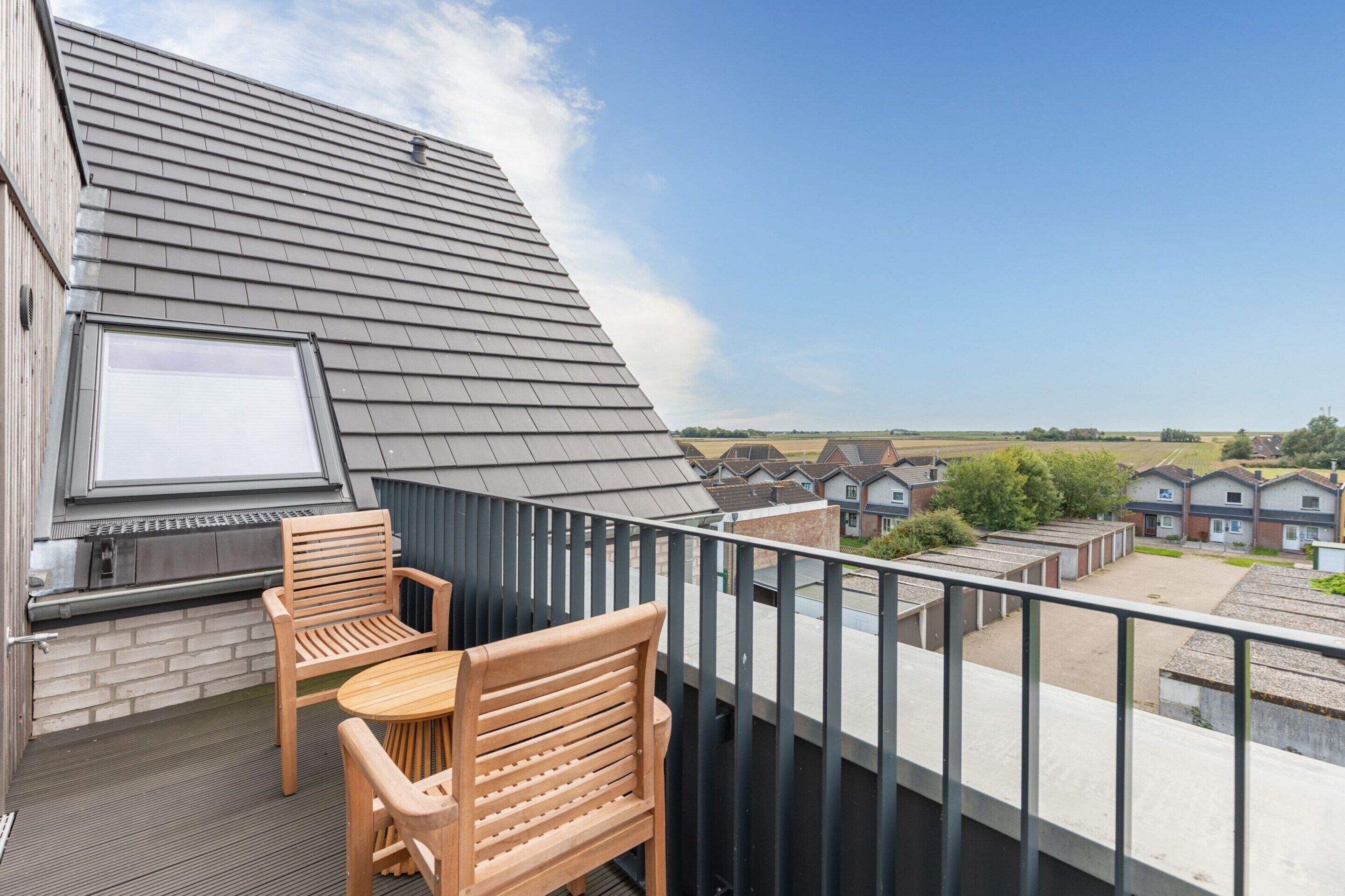 Un balcon donnant sur un quartier résidentiel, équipé d’un barbecue, de deux chaises en bois et d’une table.