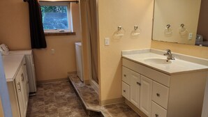 Une salle de bain avec un lavabo, un miroir et des toilettes. Il y a une fenêtre avec un rideau, et une marche qui mène à l’espace de la douche.