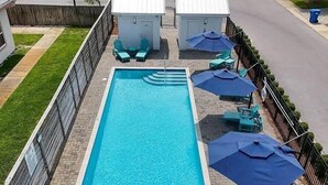 Ein rechteckiger Swimmingpool mit Fliesen umrandet, umgeben von einem Zaun, mit blauen Sonnenschirmen und Liegestühlen.