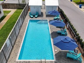 Ein rechteckiger Swimmingpool mit Fliesen umrandet, umgeben von einem Zaun, mit blauen Sonnenschirmen und Liegestühlen.