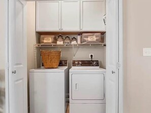 Ein Waschraum mit zwei weißen Waschmaschinen und Trocknern, einem Korbwäscheschrank und Regalen mit Taschen, auf denen 'HOME' steht.