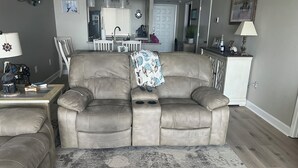 Ein Wohnzimmer mit einem beigen Sofa, einem Couchtisch und einem gemusterten Teppich. Im Hintergrund befindet sich ein Küchenbereich mit Kühlschrank, Herd und Esstisch.