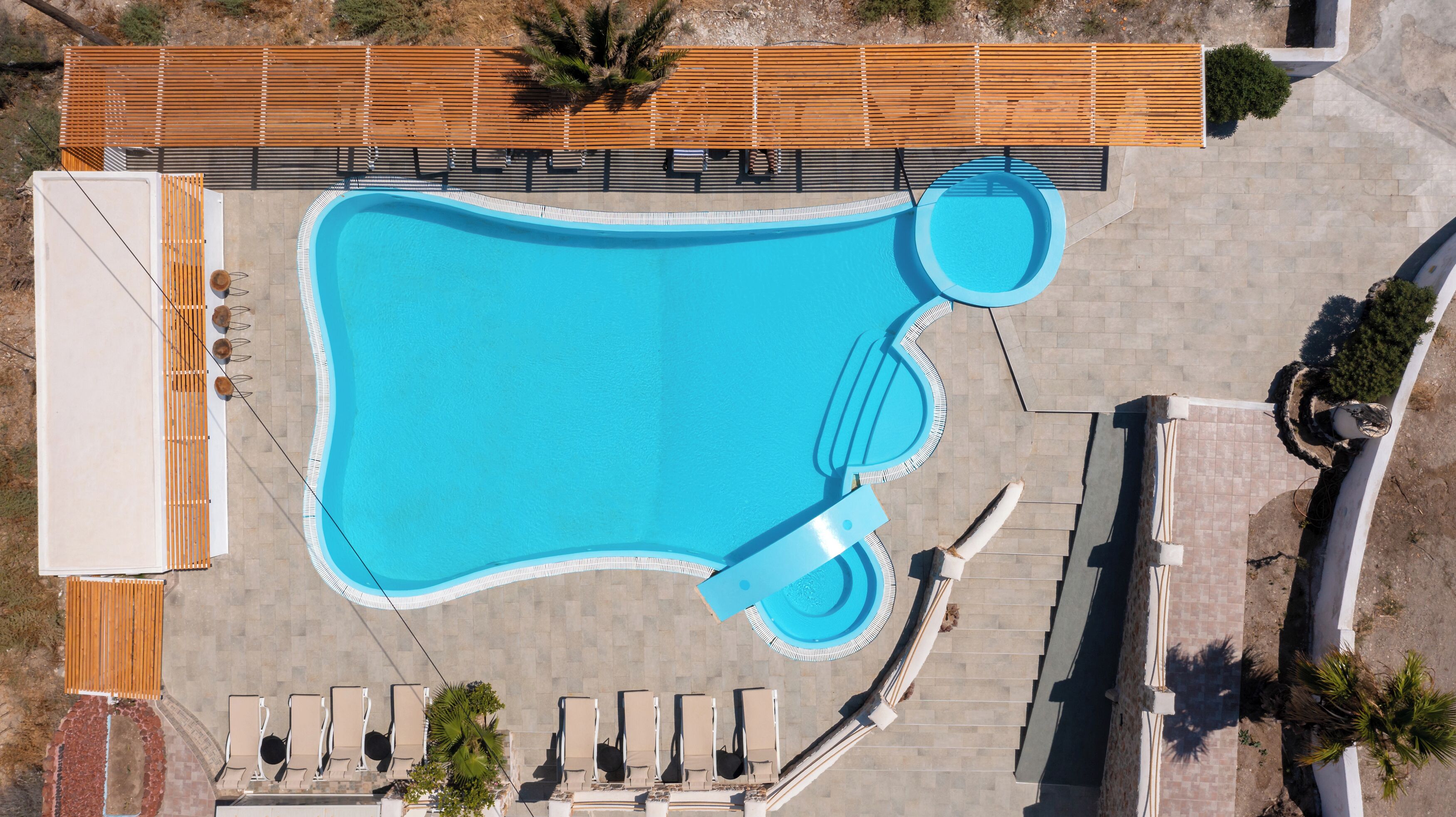 Vista aérea de una piscina con forma peculiar, rodeada de sillas de descanso y una terraza de madera.