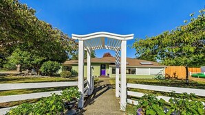 Un sendero conduce a una casa con una pérgola blanca, rodeada de plantas en macetas y árboles.