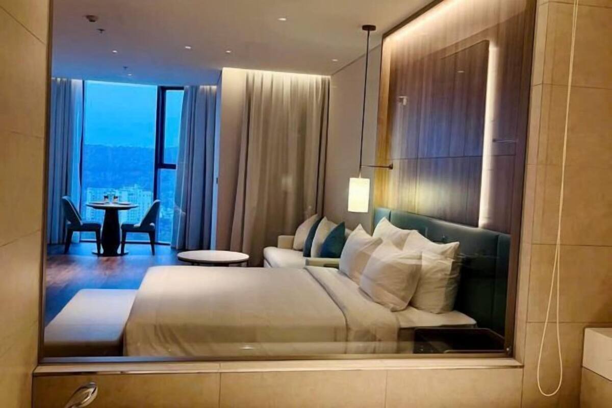 Een moderne hotelkamer met een groot bed, een klein rond tafeltje en een uitzicht op de skyline door grote ramen.