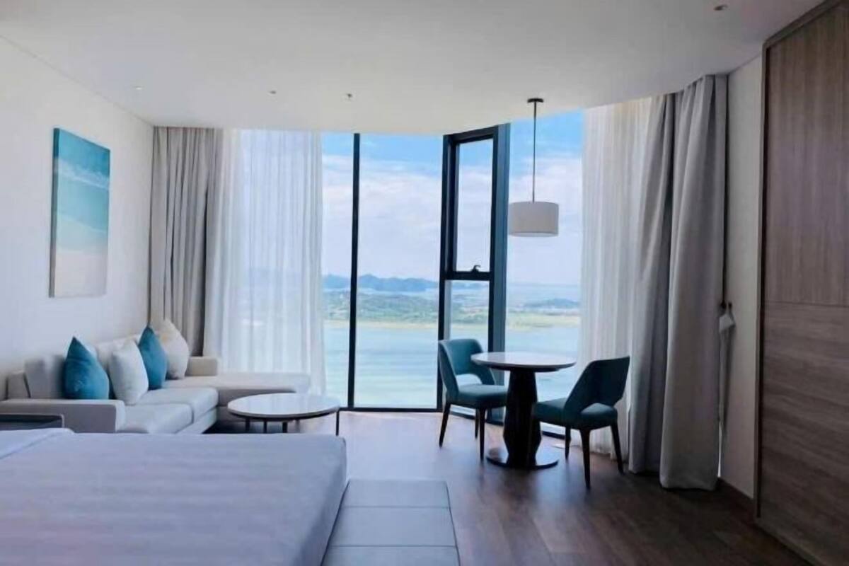 Een moderne hotelkamer met een groot bed, een bank, een eettafel en stoelen. Een groot raam biedt uitzicht op de zee en de heuvels.