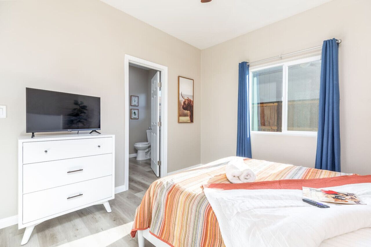 Une chambre à coucher avec un lit, un téléviseur posé sur une commode, une fenêtre avec des rideaux et une porte donnant sur une salle de bain.