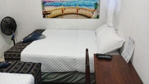 Un lit simple avec une couette blanche, une table de chevet en bois avec une télécommande, et un ventilateur fixé au mur.