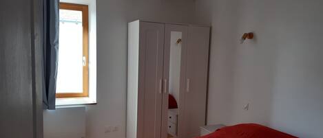 Ein Schlafzimmer mit Fenster, Kleiderschrank, Bett mit roter Decke und einem kleinen Nachttisch.