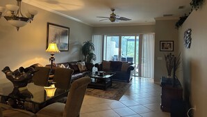 A living room with a sofa, chairs, a coffee table, a chandelier, a ceiling fan, and a large window with curtains. - Coastal Comfort in the Heart of Naples (Naples)