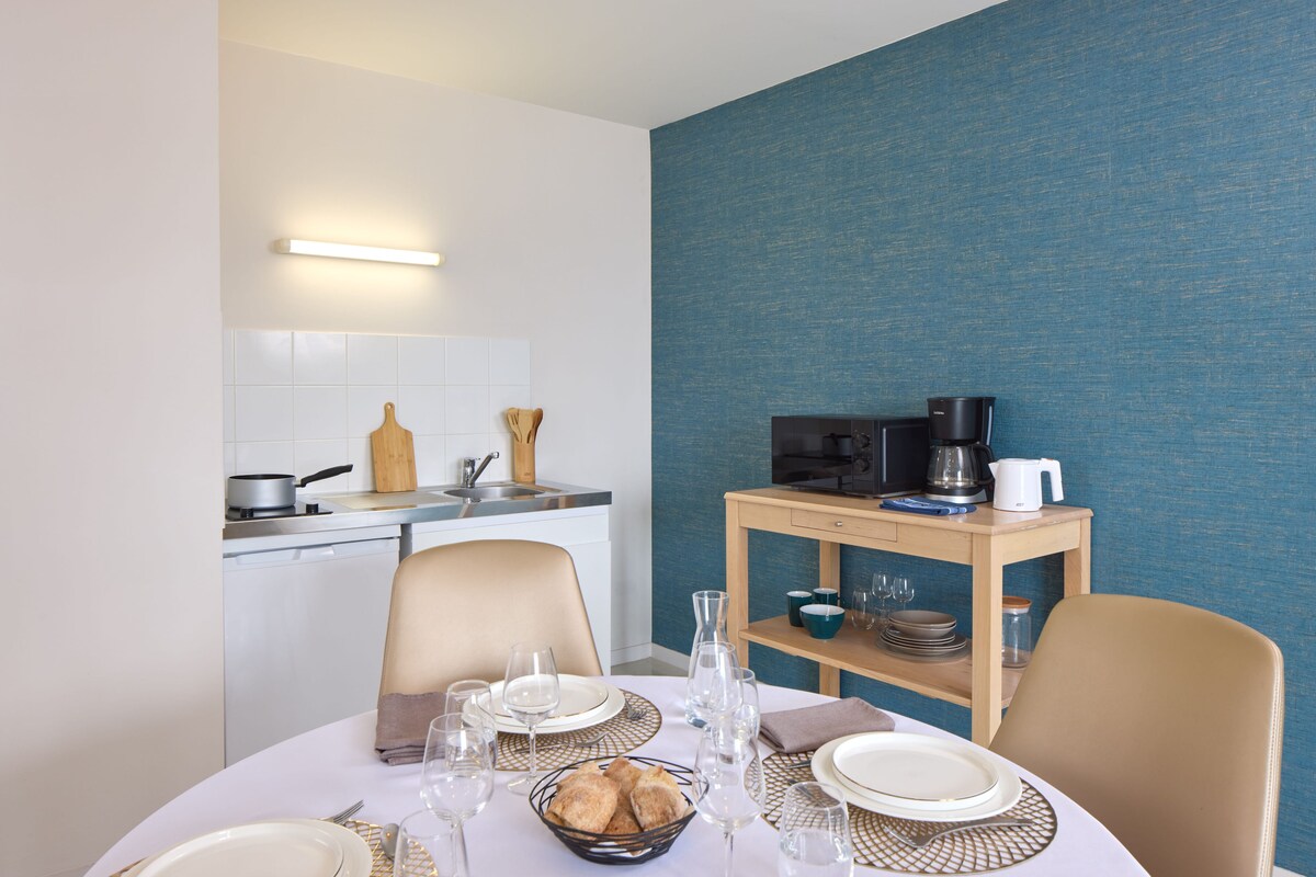 A dining area with a table set for two, a kitchen in the background, and a blue textured wall.