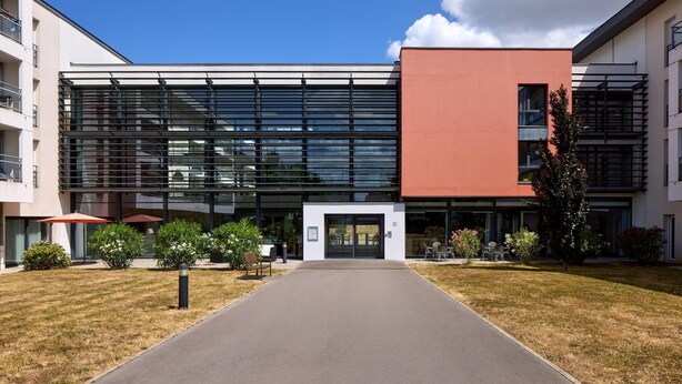 A modern building with a glass facade, a red accent wall, and a paved entrance.